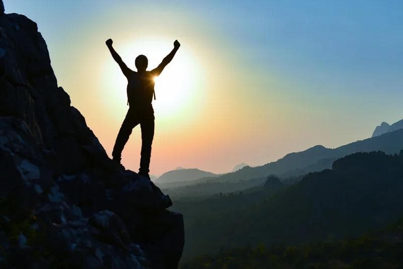 Man on mountain in front of sun holding arms up triumphantly